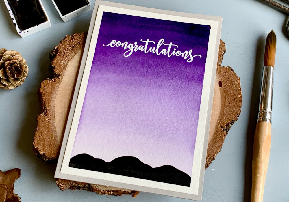 Handmade Congratulations card, with a purple ombré sky and silhouette of mountains. The greeting is heat embossed in white at the top part of the card.