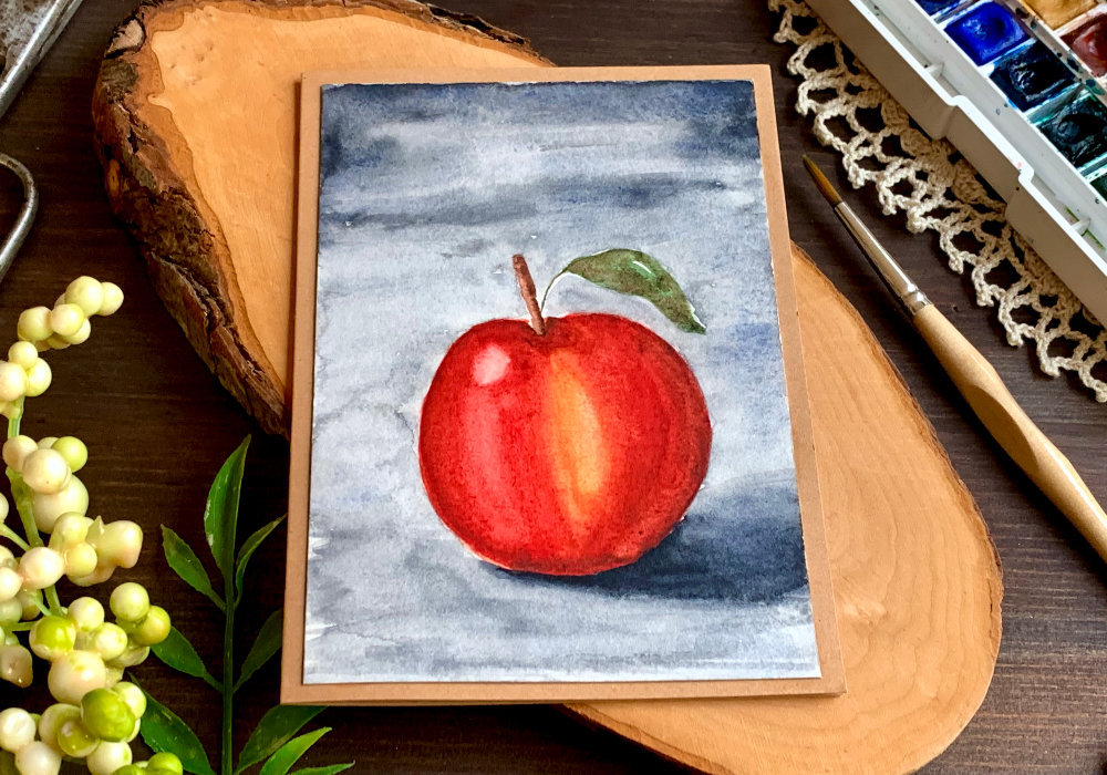 Handmade greeting card with an apple painted with watercolours. The apple is mainly red with a little bit of yellow and has one leaf. The background is dark grey, which creates a moody scene.