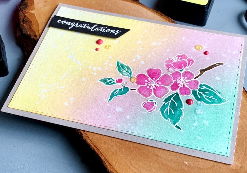 Handmade greeting card with a background in pastel colours green, pink and yellow, bended with Distress inks. Stamped cherry blossoms heat embossed in white and watercolored with the same colours. The greeting says congratulations.