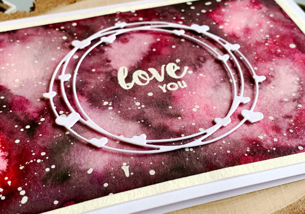 Handmade card for Valentine's day or anniversary with a pink watercolour background of a galaxy, greeting that says Love You stamped and heat embossed in the middle and white die cut with heart wreath attached in the middle of the card.