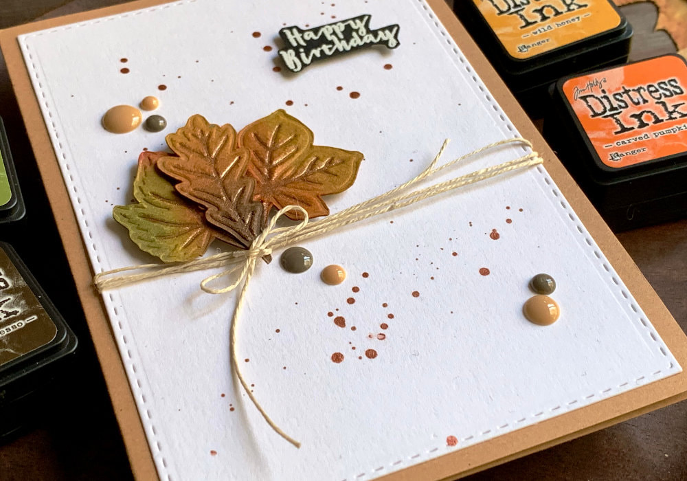 Handmade Birthday greeting card using autumnal leaves cut out form embossed background and coloured with Distress inks. The leaves are adhered onto a panel with a bakers twine attached to the background panel and the eaves and tied into a bow. The greeting says Happy Birthday and is heat embossed in white onto a black card stock.