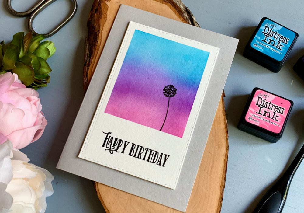 Simple Birthday card with a Polaroid shaped panel, on which is blended sunset sky in blue, purple and pink colours. The sky is created using Distress inks Mermaid Lagoon, Wilted Violet and Picked Raspberry. On the background is stamped single black flower and below it is stamped Happy Birthday greeting.