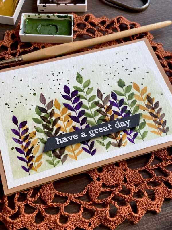 Handmade greeting card with hand-painted watercolour leaves along the bottom edge with straight stems, in the colours dark yellow, green, purple and brown. The greeting says Have A Great Day and is stamped and heat embossed in white on a black card stock, cut into a banner and adhered in the lower part of the card.