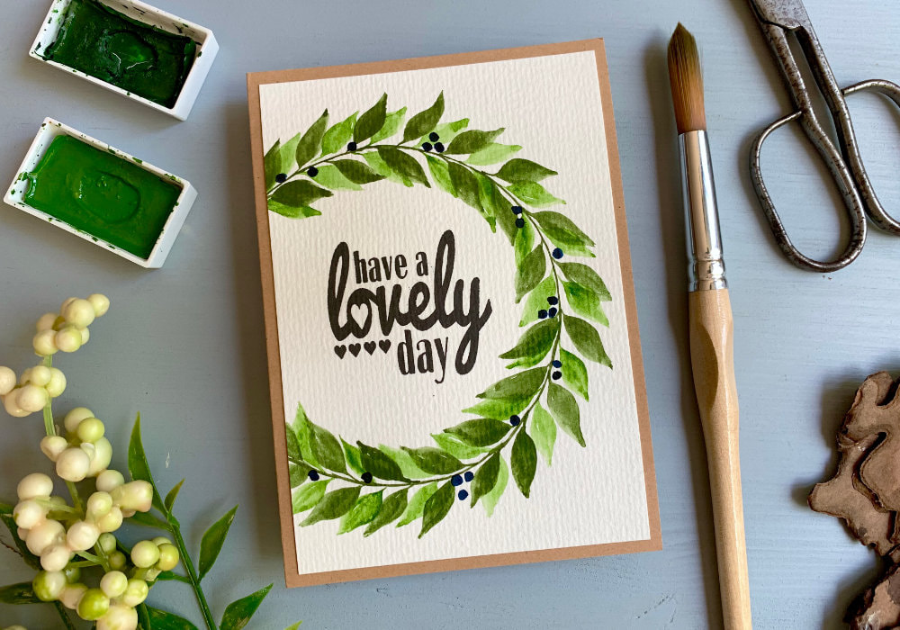 Watercolour painting of a wreath in two shades of green and blue berries. One of the paintings is made into a greeting card with a stamped greeting in the middle saying Have A Lovely Day. The other paining is placed into a picture frame and in the middle has a stamped quote saying  Live Every Moment, Laugh Every Day, Love Beyond Words.