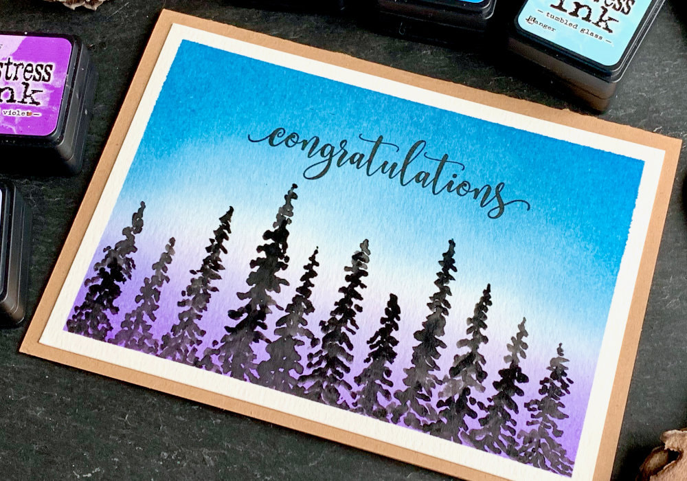 Handmade card with a sunset/sunrise background created with Distress inks and black silhouettes of pine trees at the bottom painted with watercolours.