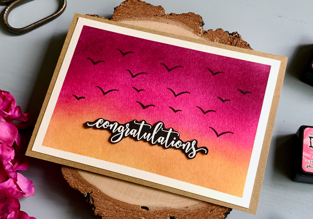 Handmade congratulation card with a sky background at sunset created using Distress inks in colours purple, pink and yellow and stamped bird silhouettes flying in the sky.