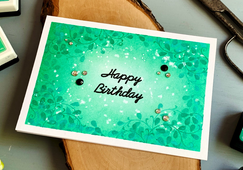 Handmade Birthday card with tone on tone background made by blending two shades of Distress inks in the same colour - turquoise and stamping solid leaves with turquoise dye inks to create a monochrome background. The greeting is stamped with a black ink in the middle of the card and says Happy Birthday.