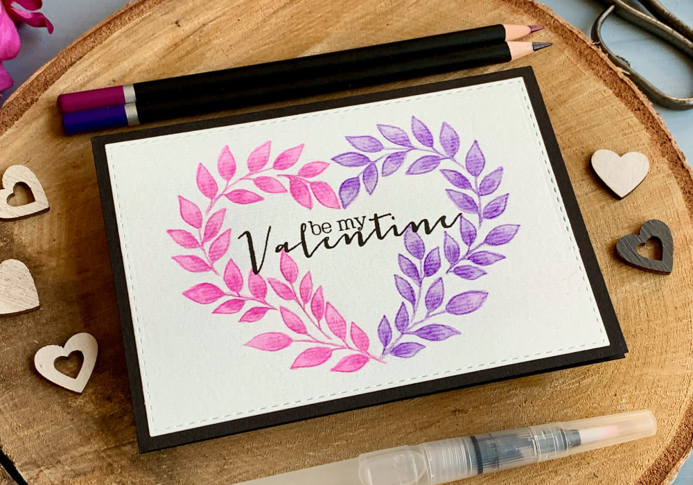 DIY greeting card for Valentine's day with wreath in a shape of a heart hand drawn using watercolour pencils, with one half of the heart painted in pink and the other purple and greeting saying Be My Valentine stamped in black in the middle and black card base.