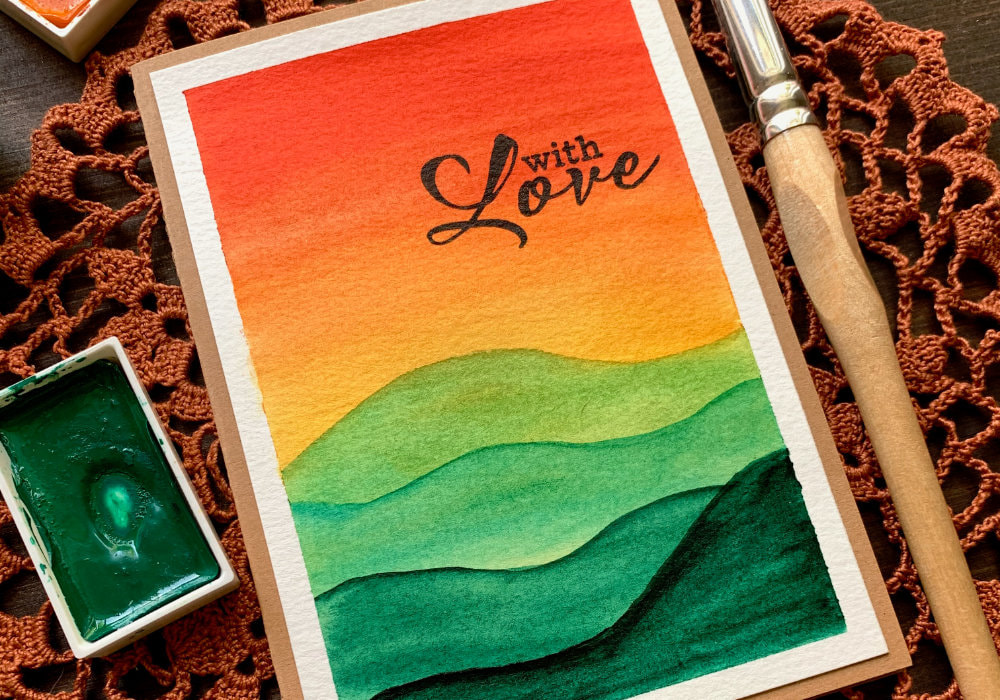 DIY card with hand-painted autumnal landscape. Watercolour unset sky going from red to orange and yellow and green mountains. The greeting is stamped with black ink at the top right corner and says 