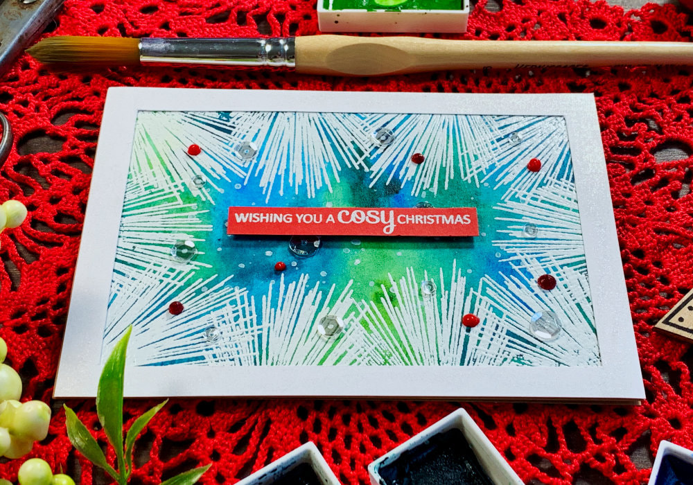 Handmade Christmas card with a green and blue watercolour background and white heat embossed pine needles along the edge of the panel creating a frame. Greeting says 