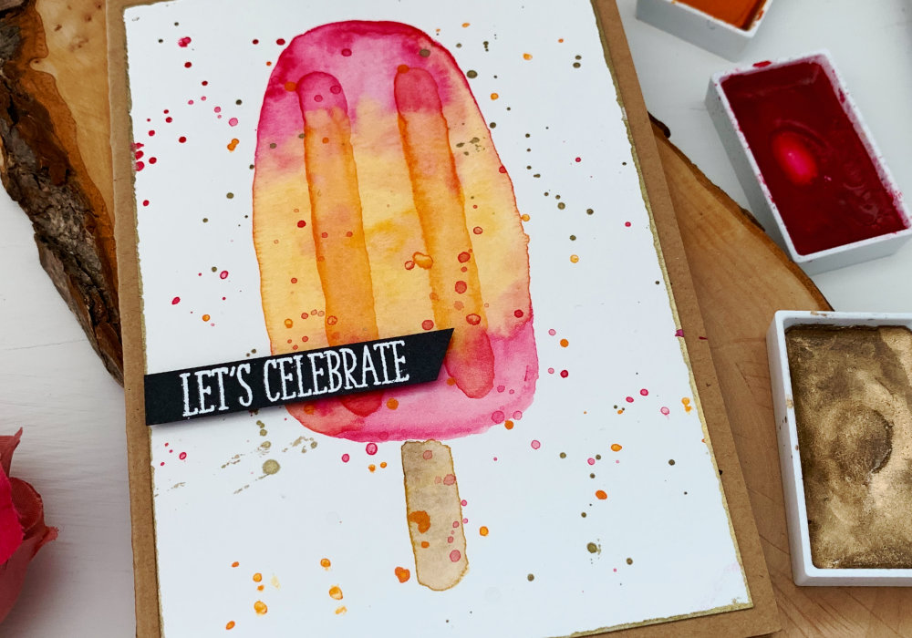 Handmade Birthday card with a watercolour ice lolly / popsicle, pink on the top and bottom and orange in the middle. The greeting says Let's Celebrate and it's stamped on a black card stock, heat embossed in white and cut it out into a banner.