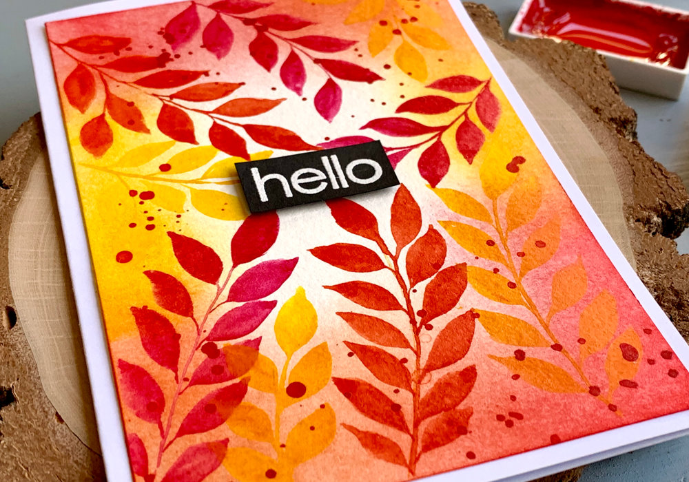 Handmade greeting card saying Hello with a background with watercolour leaves painted in yellow, pink and orange along the edges of the card stock. With the help of Distress inks in matching colours and blended along the edges it brings the whole background together. 