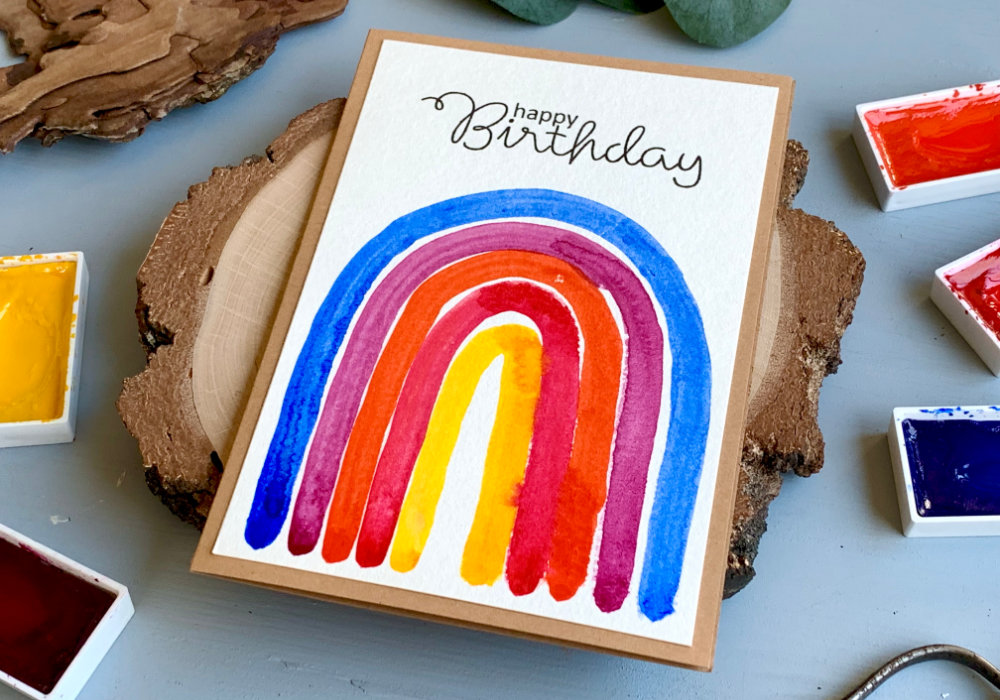 DIY Birthday card with a hand painted rainbow in vibrant colours - blue, purple, orange, pink and yellow. The greeting says Happy Birthday and is stamp in black ink above the rainbow. The watercolour panel is adhered on an A6 card base made out of a craft card stock.