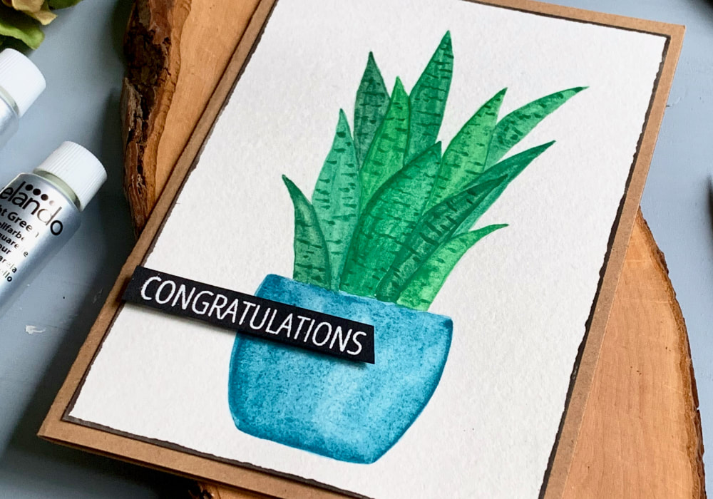 Handmade Congratulations card with a hand-drawn and painted snake plant using watercolours. The pot of the plant is blue and the plant is bright green. A greeting that says Congratulations is stamped and heat embossed in white on a black banner and adhered on top of the watercolour panel. 