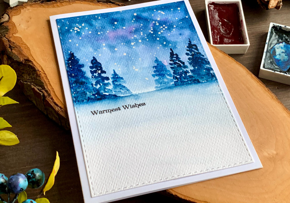 Handmade Christmas card with a watercolour winter forest scene at night. The forest is painted in the top half of the card and the bottom is left white. The greeting says Warmest Wishes and is stamped below the trees.