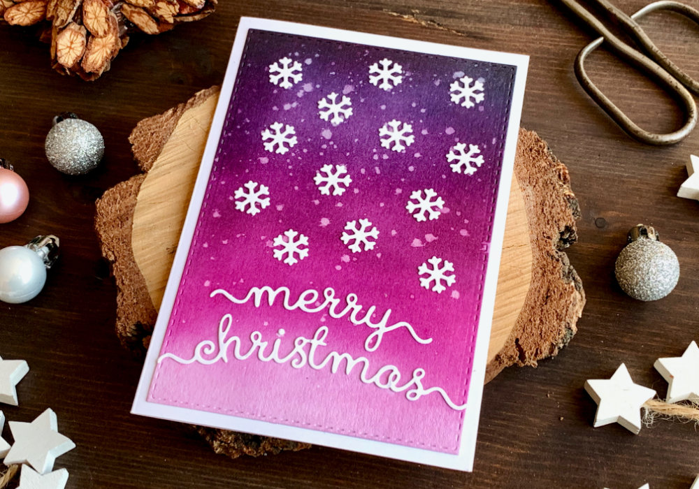 Handmade Christmas card with a pink and purple background created by blending the Distress inks  Black Soot, Faded Jeans, Seedless Preserves, Picked Raspberry, and adhering white die-cut snowflakes as well as a Merry Christmas greeting over the panel.