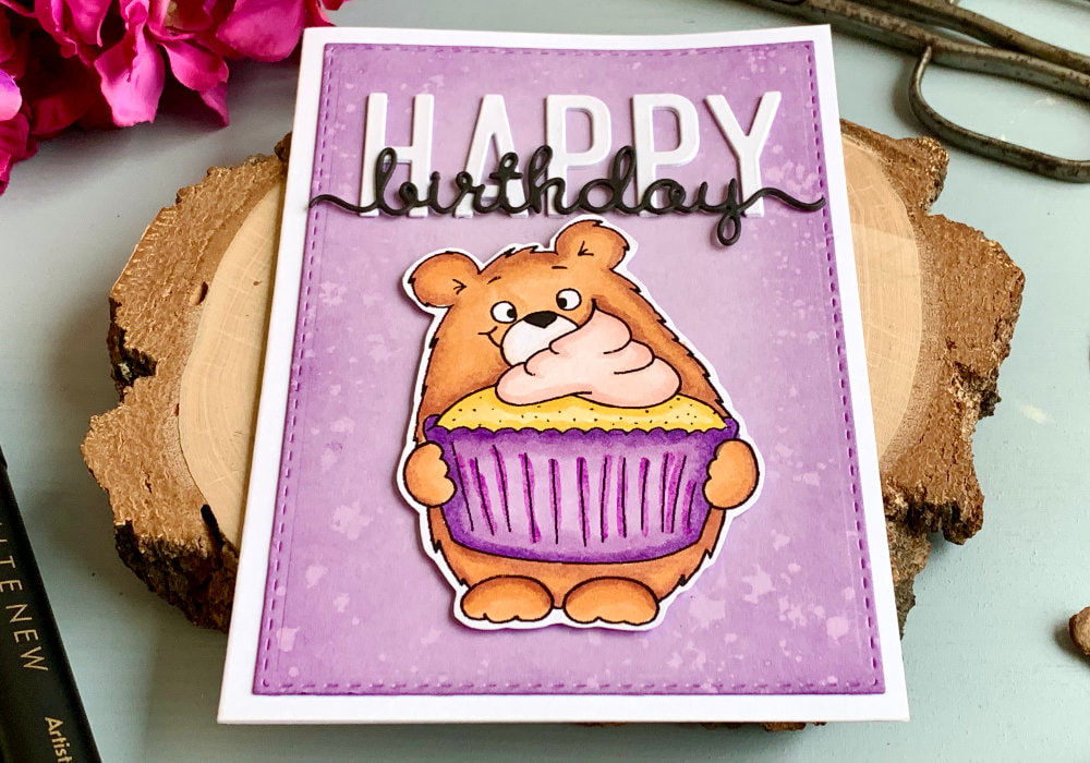 Handmade Birthday card with a bear holding a cupcake, coloured with alcohol markers. The bear is brown and cupcake liner is purple. The bear is cut out and adhered on top of a lilac panel with a bold Happy Birthday sentiment in white and black letters.