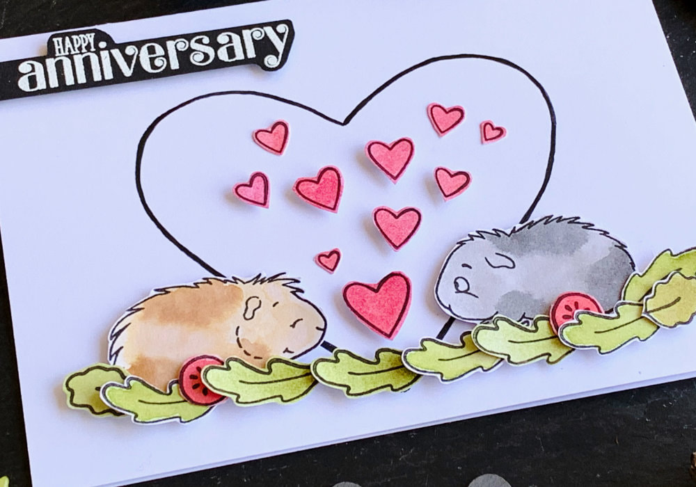 Handmade Anniversary card with stamped two guinea pigs facing each other, one grey, one brown, standing in salad leafs and tomatoes. In between them are pink hearts and in the background is a big black outline of a heart. 