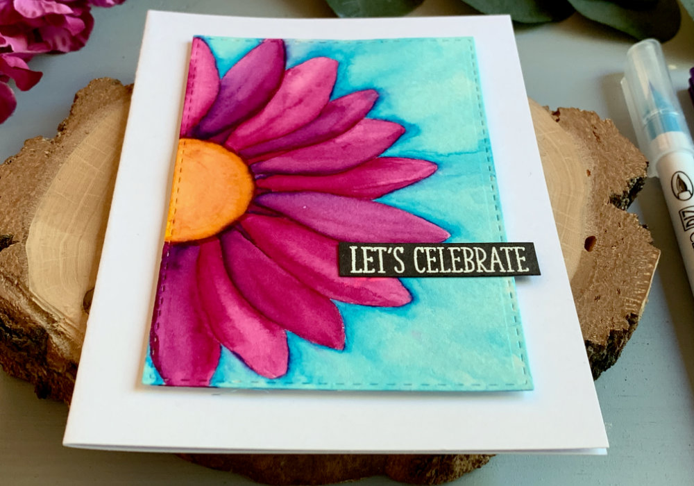 A handmade Birthday card with a big bold daisy flower with bright pink petals and blue background, coloured with waterbased markers and adhered on a white card base. The greeting says Let's Celebrate.