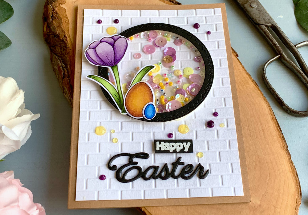 Handmade greeting card for Easter with a circle shaker element filled with sequins and beads, crocus flower and eggs on top of the shaker, a white background panel with a brick pattern created using an embossing folder and the word Easter die-cut from a black card stock and the word happy stamped and heat embossed in white and cut into a banner. All adhered on top of a card base made out of a craft card stock.