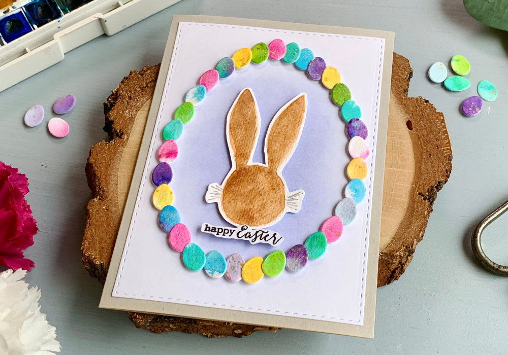 Handmade Easter greeting with a wreath created with tiny egg dies, watercolour backgrounds in various colours and hand-drawn, watercolour head of a bunny in the center of the wreath.
