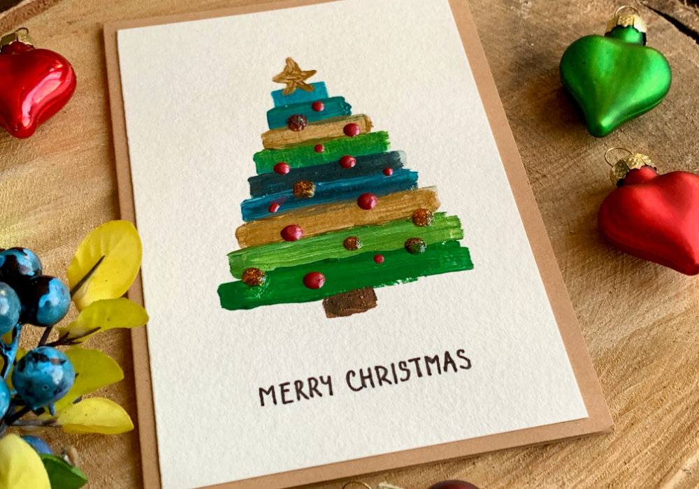 Handmade Christmas cards with a Christmas tree one with watercolours and one acrylic paints, painting simple stripes to create a triangle like shape, using green, blue and gold paints.