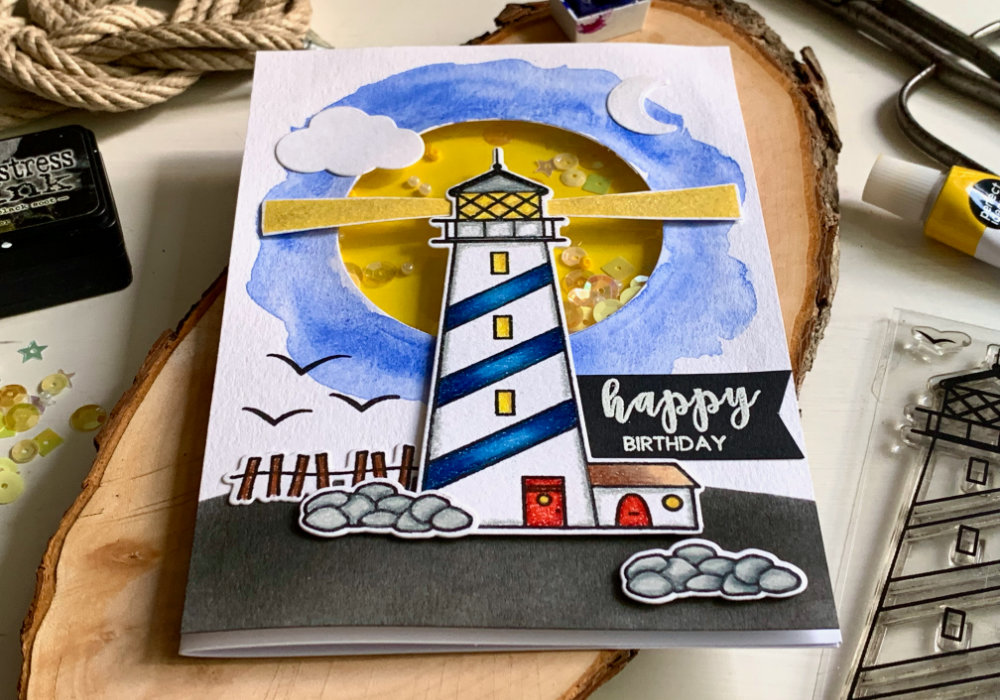 Handmade Birthday shaker card with a lighthouse at night on a hill, placed over a shaker window with a yellow background and sequins and beads. 