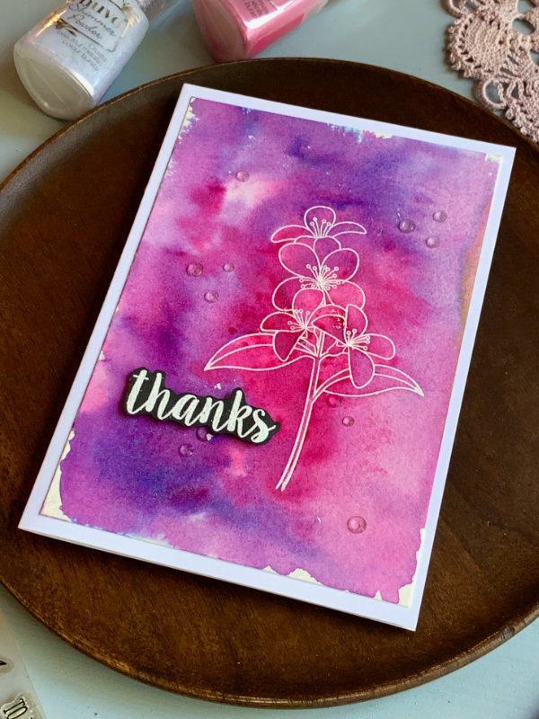 Handmade card with a pink and purple background created with the Nuvo Shimmer Powders. On top of the card is stamped and heat embossed white flower. The greeting is  heat embossed in white on black card stock and it says Thanks. 
