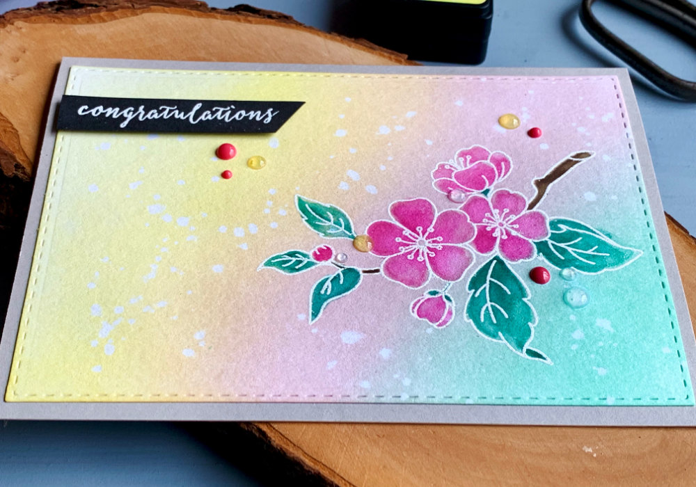 Handmade greeting card with a background in pastel colours green, pink and yellow, bended with Distress inks. Stamped cherry blossoms heat embossed in white and watercolored with the same colours. The greeting says congratulations.