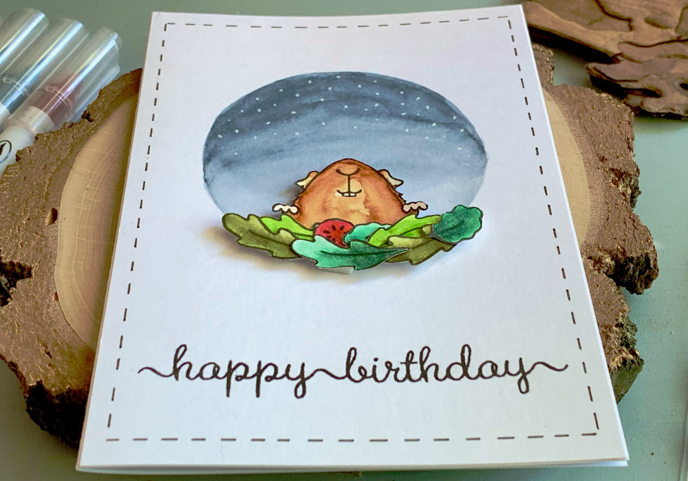 Handmade Happy Birthday card with a stamped image of very happy guinea pit peaking from behind of stacked salad leaves. The guinea pig is at the bottom of a painted circle in the colours of a night sky with stars.