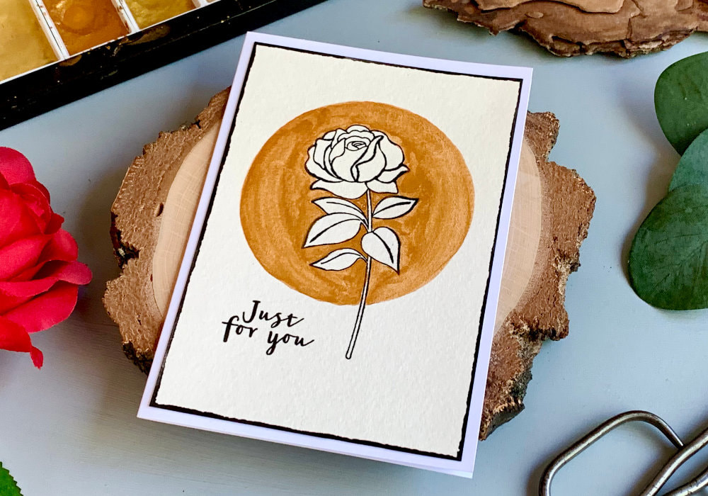 Handmade greeting card not only for Birthday, with a stamped rose using  a black ink. The background around the flower is painted with gold watercolours in a circle. The rose is unpainted. The greeting says Just For You.