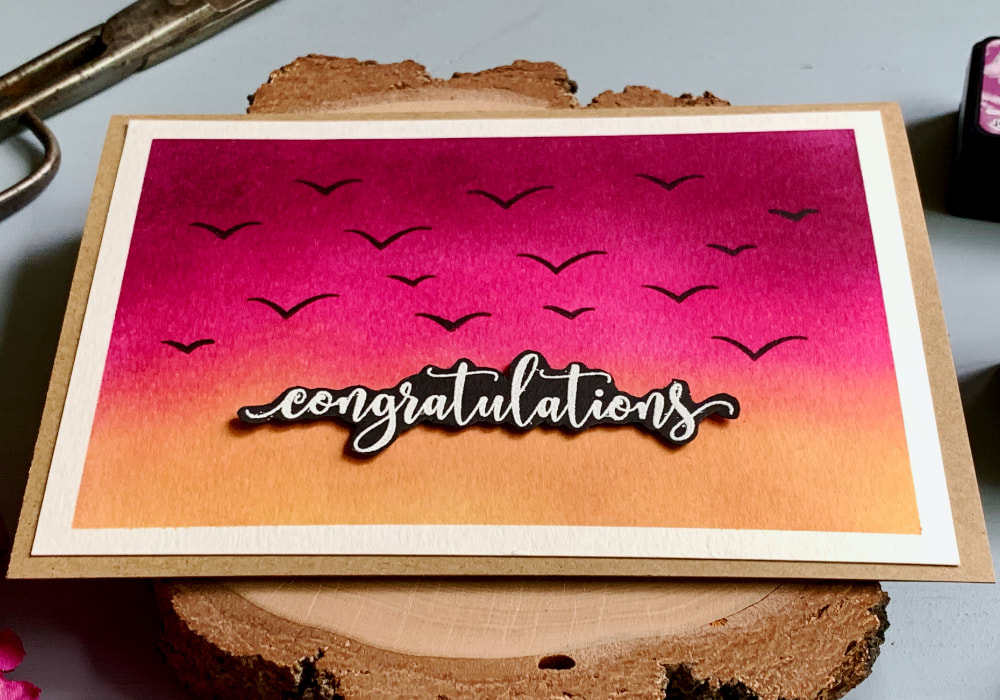 Handmade congratulation card with a sky background at sunset created using Distress inks in colours purple, pink and yellow and stamped bird silhouettes flying in the sky.