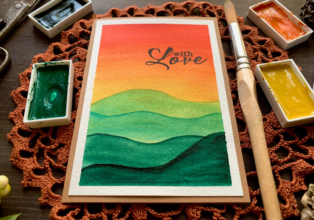 DIY card with hand-painted autumnal landscape. Watercolour unset sky going from red to orange and yellow and green mountains. The greeting is stamped with black ink at the top right corner and says 