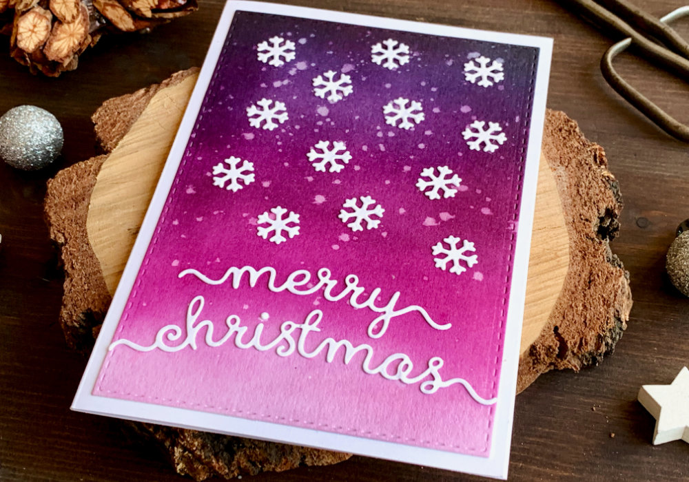 Handmade Christmas card with a pink and purple background created by blending the Distress inks  Black Soot, Faded Jeans, Seedless Preserves, Picked Raspberry, and adhering white die-cut snowflakes as well as a Merry Christmas greeting over the panel.