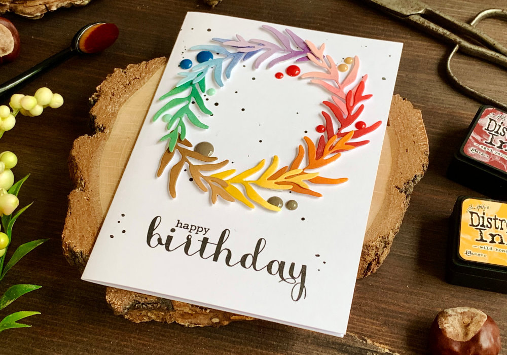 Handmade Birthday greeting card with a wreath made using a leaf die and adhered onto a card base in a circle. The wreath is assembled in rainbow colours, each branch is coloured using Distress inks. There is a watercolour splatter as well as some enamel dots to add interest. And the greeting say Happy Birthday and is stamp in black below the wreath.