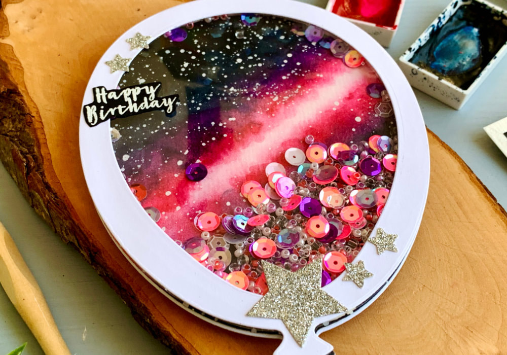 Handmade Birthday shaker card in a shape of a ballon, filled with beads and sequins in different pink and purple colours. The background of the card is painting of a pink galaxy painted with watercolours.
