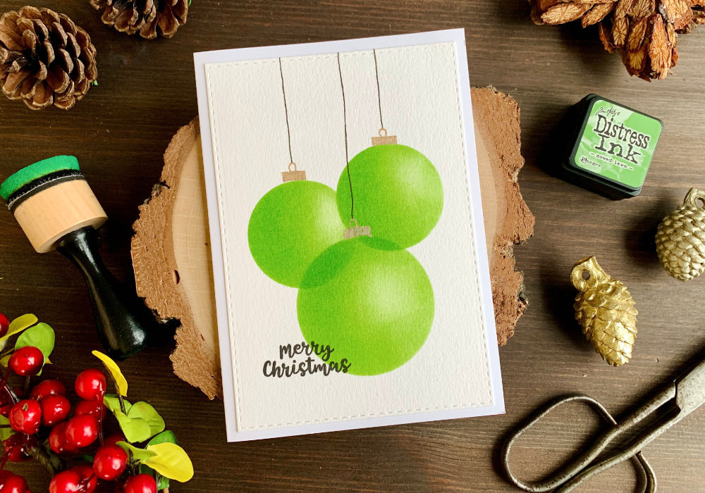 Handmade Christmas card with baubles created with Distress inks and a (DIY) circle stencil. Three overlapping green baubles, with golden tops with loops and black strings. The greeting says Merry Christmas.