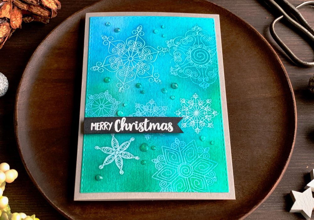 Simple handmade Christmas card with a blue-turquoise background created by using different shades of green and blue inks and snowflakes that were heat embossed using a clear embossing powder to keep the colour of the layer below. The greeting says Merry Christmas and is stamped and heat embossed in white on a black card stock, cut into a banner and adhered with a foam tape on top of the snowflake background.