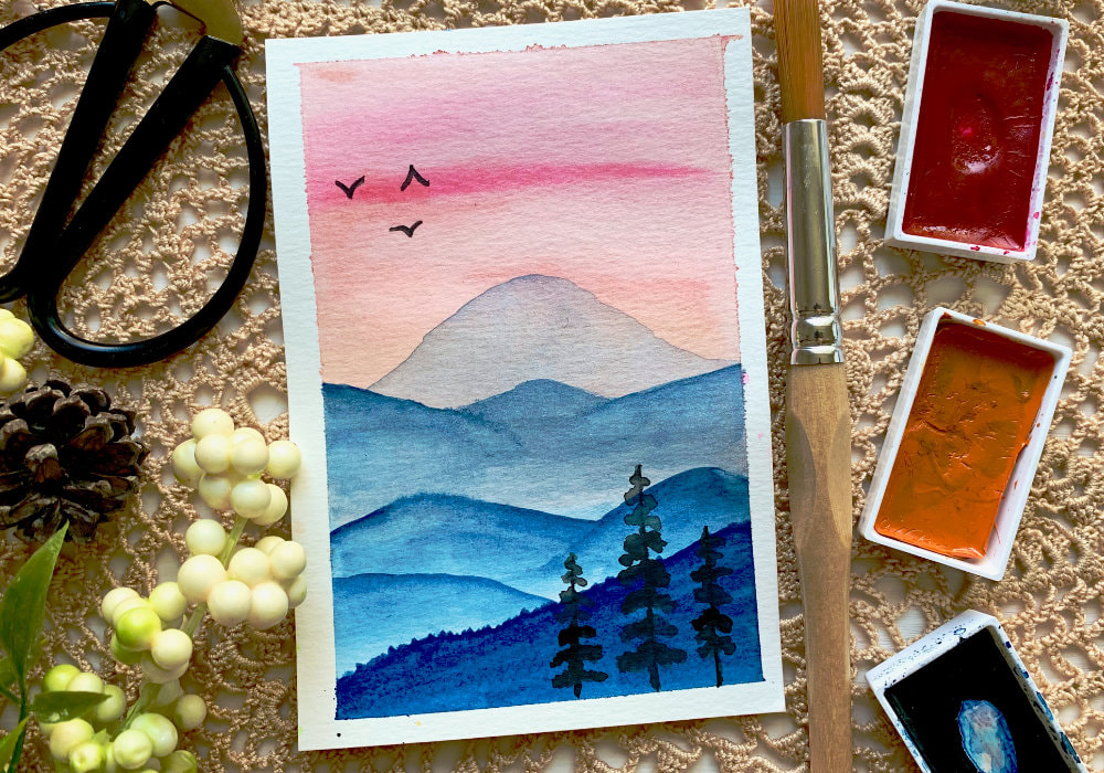 Handmade card with watercolour landscape with pink sky and blue mountains with trees and birds in the sky..