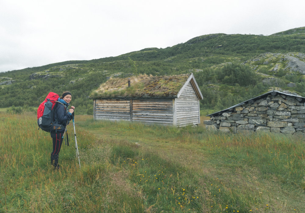 Hiking in Hardangervidda in Norway, photo of me with my red backpack next to a wooden house.