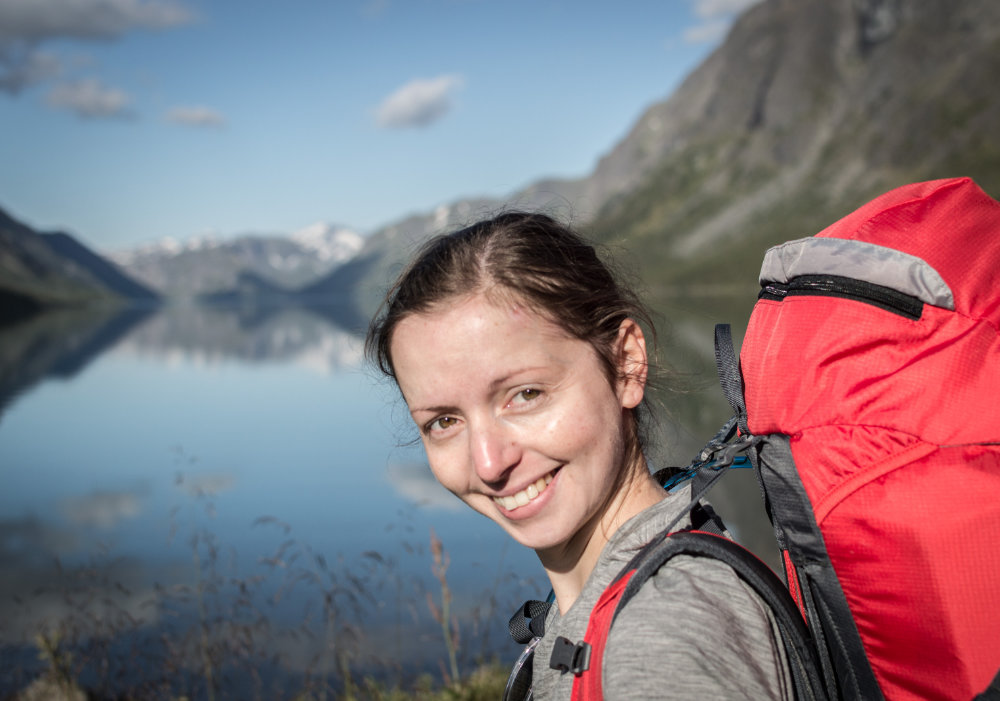 Photo of myself, with a red hiking backpack on my back and the lake Gjende and vast Norwegien mountains in the background.