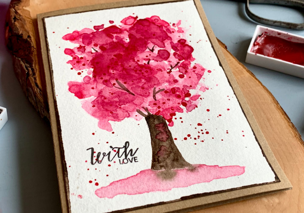 DIY greeting card with a hand-painted cherry tree using pink watercolours and the ink smooshing technique. The greeting says With Love, stamped with a black ink. 
