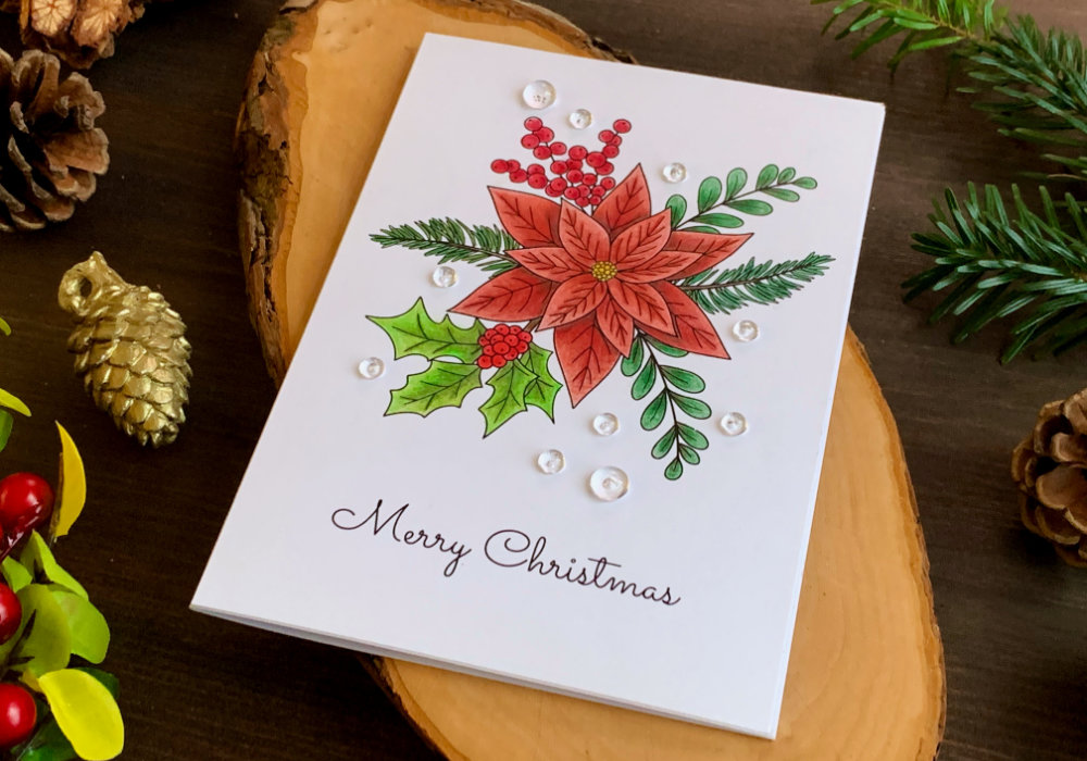 Handmade Christmas card with an image of poinsettia bouquet including a holly and berry and fir tree branches. The image is coloured with the Faber-Castell Polychromos colouring pencils using reds and greens.