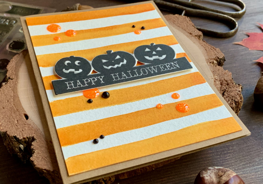 Simple handmade Halloween card with a background with orange stripes painted with watercolours and a greeting with pumpkins and a Happy Halloween sentiment, stamped in black.
