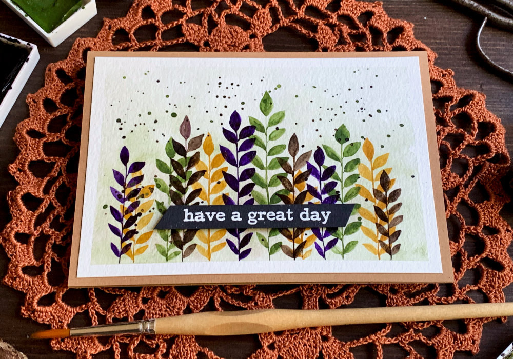 Handmade greeting card with hand-painted watercolour leaves along the bottom edge with straight stems, in the colours dark yellow, green, purple and brown. The greeting says Have A Great Day and is stamped and heat embossed in white on a black card stock, cut into a banner and adhered in the lower part of the card.