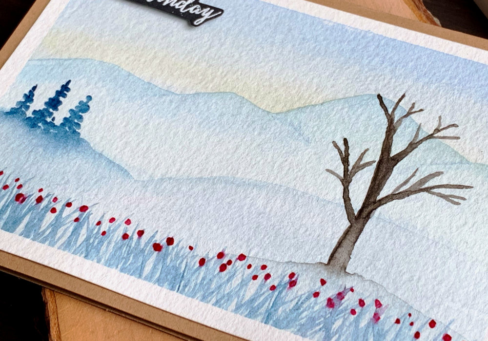 Handmade card with a winter watercolour landscape. With snowy mountains in the background, small pine trees in the middle and a big tree, without leaves in the front.
