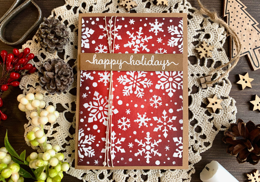Make a handmade Christmas card with a red Distress ink blending using the inks Ground Espresso, Aged Mahogany, Barn Door and Candied Apple. Covered with snoflakes stamped with a stencil and heat embossed in white.