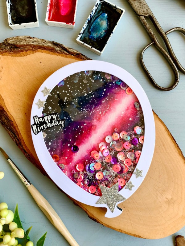Handmade Birthday shaker card in a shape of a ballon, filled with beads and sequins in different pink and purple colours. The background of the card is painting of a pink galaxy painted with watercolours.