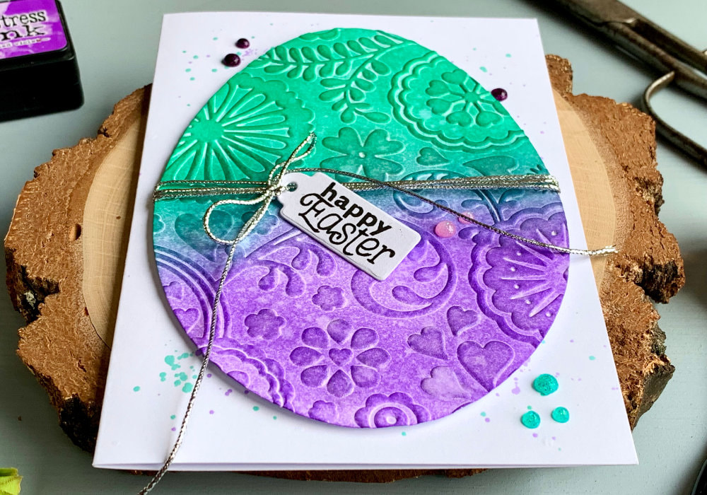Handmade card for Easter with a big die-cut egg that is embossed with an embossing folder and coloured using Distress inks Lucky Clover - green and Wilted Violet - purple. The greeting says Happy Easter and is stamped onto a tag which is attached to a silver string that is attached to the egg. The egg is adhered on top of a white card base.