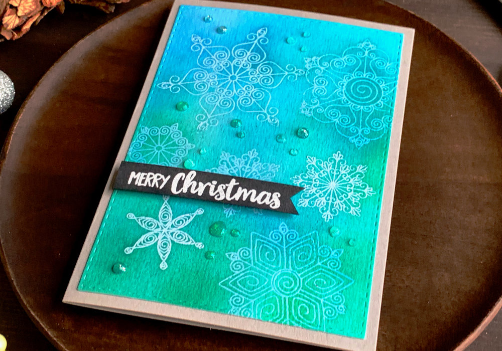 Simple handmade Christmas card with a blue-turquoise background created by using different shades of green and blue inks and snowflakes that were heat embossed using a clear embossing powder to keep the colour of the layer below. The greeting says Merry Christmas and is stamped and heat embossed in white on a black card stock, cut into a banner and adhered with a foam tape on top of the snowflake background.